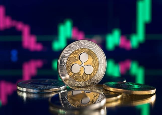 Eine Ripple-Münze (XRP) steht aufrecht zwischen anderen Münzen, mit einem digitalen Candlestick-Chart mit grünen und rosa Balken im unscharfen Hintergrund, was auf einen aktiven XRP-Futures-Handel auf Plattformen wie Coinbase hindeutet.