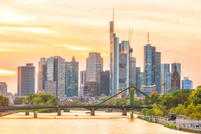 Eine Skyline mit modernen Wolkenkratzern – Sitz führender Krypto-Finanzunternehmen – und einer grünen Brücke über einen Fluss bei Sonnenuntergang, mit Bäumen am Flussufer.